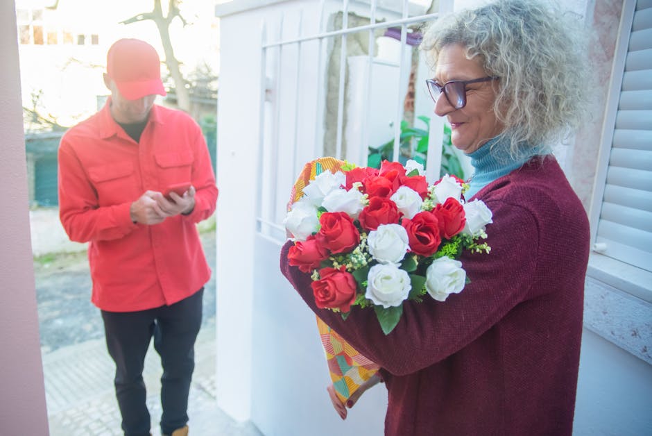 A woman with curly gray hair, wearing glasses, a maroon sweater, and a blue turtleneck, is holding a vibrant bouquet of fresh red and white roses wrapped in decorative paper near a doorway, with sunli