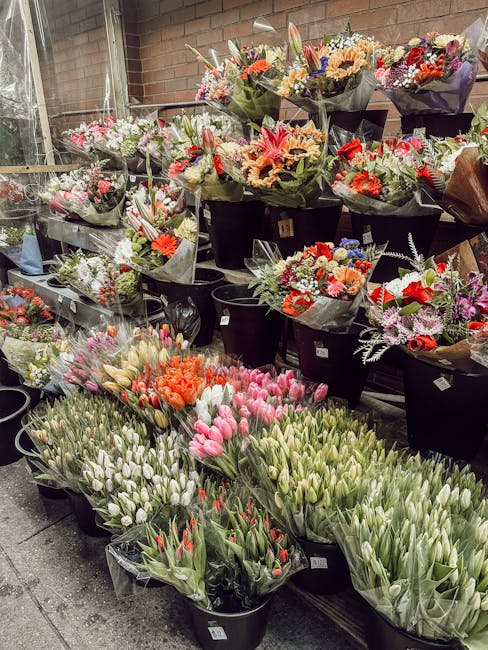 An assortment of fresh flower bouquets arranged in black buckets and placed on shelves and the floor, featuring a variety of colorful flowers including pink, white, orange, and purple blooms. The bouq