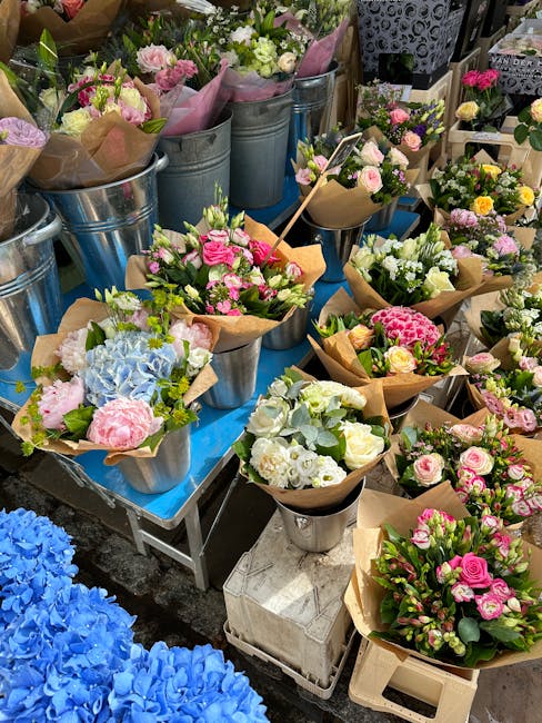 A display of fresh flower bouquets arranged in metal buckets and wrapped in brown paper on a market stall. The selection includes pink roses, white lisianthus, blue hydrangeas, and various greenery, a
