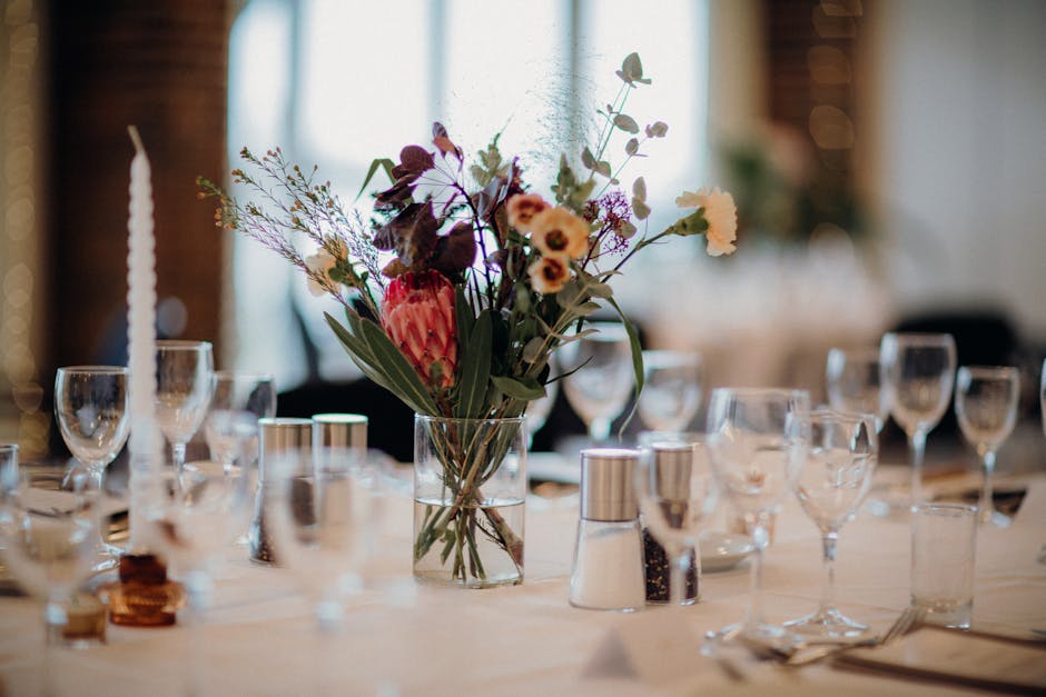 A floral arrangement featuring a variety of fresh flowers, including pink protea, dark purple foliage, and small white and purple blossoms, styled in a clear glass vase placed at the center of a dinin