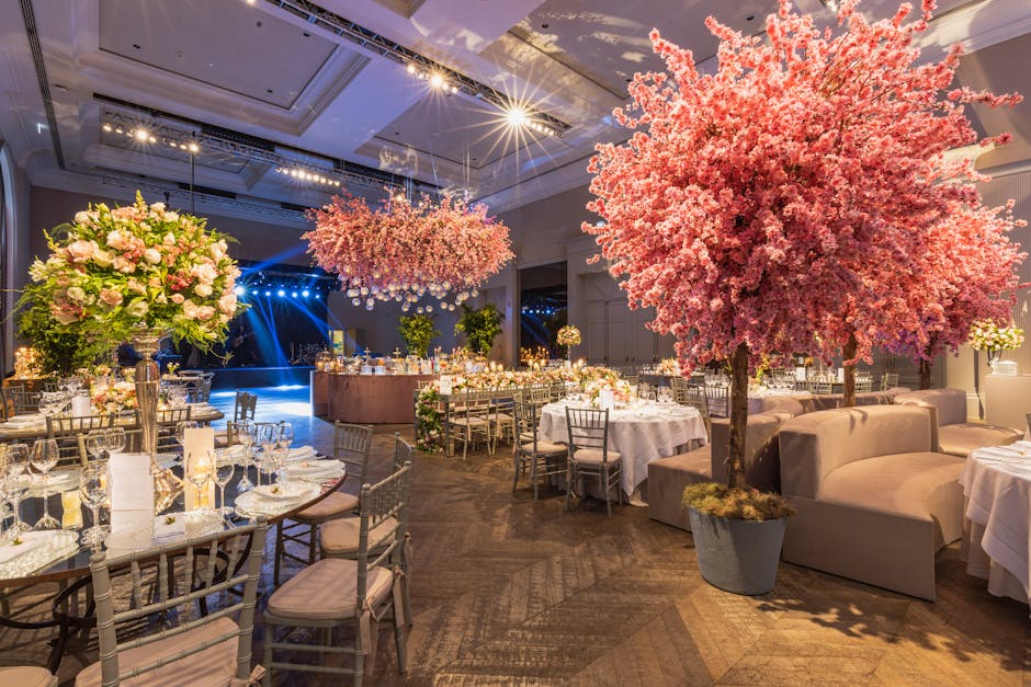 An elegant banquet hall decorated for a wedding, featuring large, vibrant pink cherry blossom trees in grey pots positioned among round tables draped with white tablecloths. Each table is set with whi
