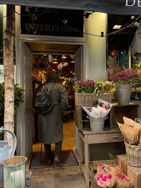 A person with a dark coat and a black backpack stands at the entrance of a floral shop, [floristhackney.co.uk], on Mare Street Hackney. Outside the shop, there are baskets and pots filled with fresh p