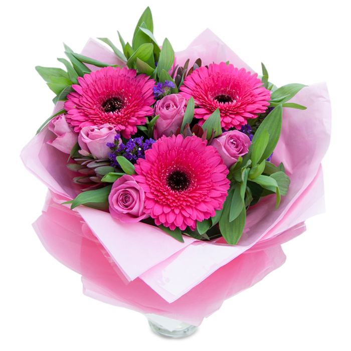 I picture this bouquet as a small, joyful scene captured in flowers: three wide-faced Gerbera daisies in assertive fuchsia take the centre stage, each petal catching the light and directing your gaze toward their deep, dark centres. Complementing them are soft, dusty lavender-pink roses whose petals are still forming that intimate spiralled cup, offering a softer, more romantic counterpoint to the gerberas' punch. Scattered between these principal blooms are delicate sprigs of purple statice that float like confetti, introducing texture and a hint of meadow-like spontaneity. The green backdrop reads as garden-fresh-broad salal or eucalyptus leaves with a slightly glossy surface that gives the bouquet structure and a crispness reminiscent of morning dew. The hand-tied arrangement is presented in layered wrap: the inner pale pink tissue is matte and protective, while an outer translucent fuchsia-pink cellophane with a subtle crinkle reflects light and unifies the palette. The very bottom shows the rim of a clear glass vase, signaling the bouquet is meant for immediate display. This arrangement feels intimate and celebratory, ideal for anniversaries, birthdays, or simply to lift someone's spirits. I can easily see it brightening a narrow studio near Hackney Wick, placed on a kitchen table after a visit to Chatsworth Road or left as a thoughtful surprise for a neighbour on Graham Road, its colours designed to bring warmth and cheer to a Hackney home.